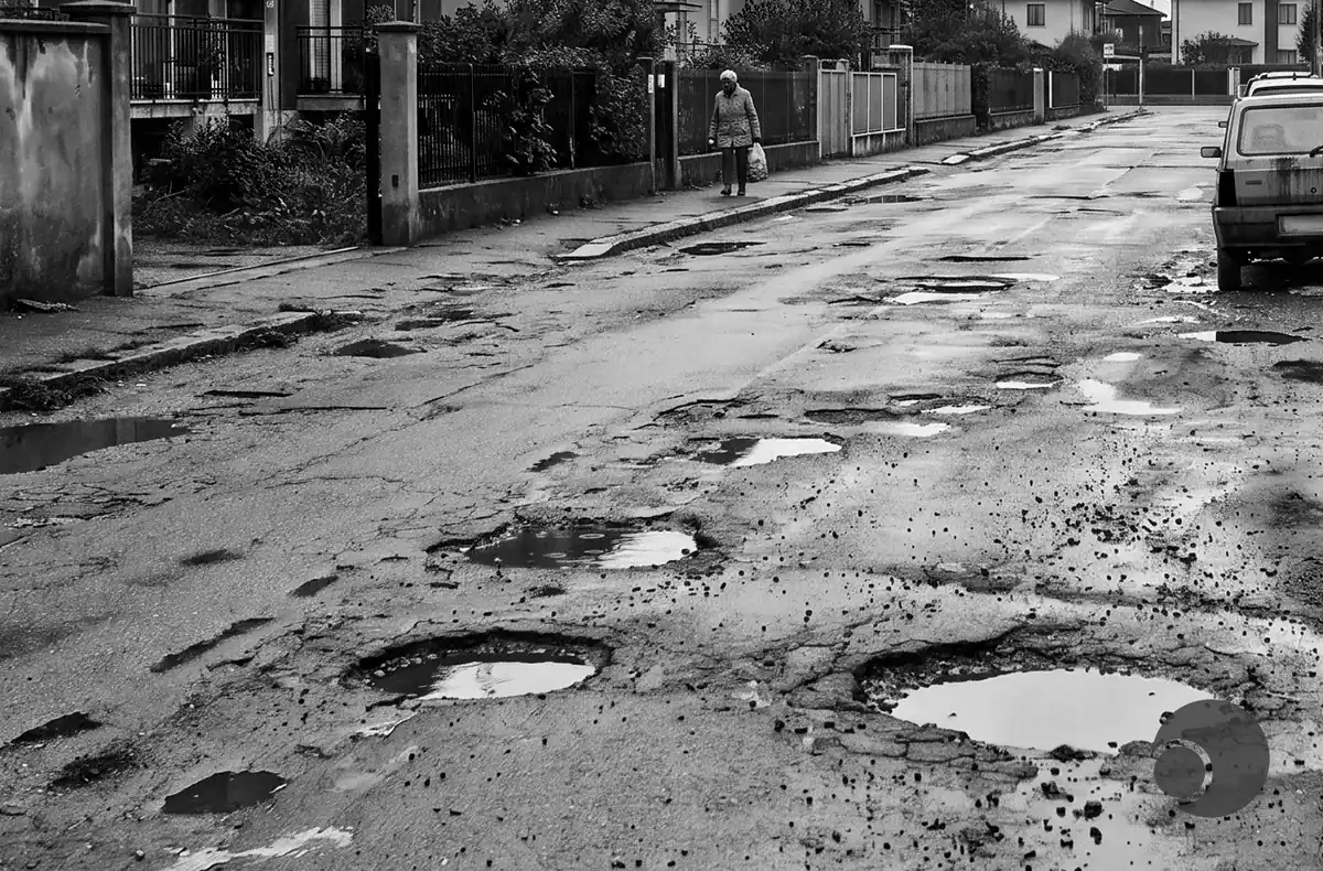 Residential street with numerous potholes and puddles; a person walks on the sidewalk carrying a bag.