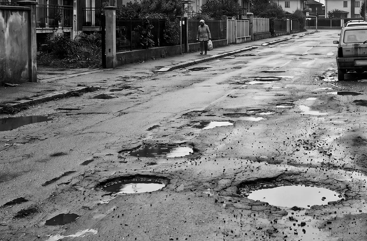 Residential street with numerous potholes and puddles; a person walks on the sidewalk carrying a bag.